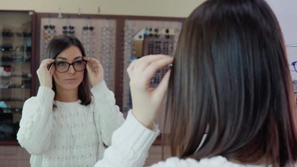 Young Brunette with Blue Eyes in a Cozy White Sweater Trying on Glasses in Front of a Mirror in an