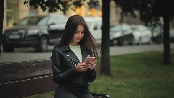 A Young Woman is Sitting on a Bench and Texting on Her Smartphone