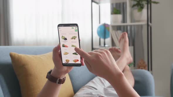 Man Lying On Couch Using Smartphone Buys In Internet Shop. Shop Vegetables In Grocery Online Store