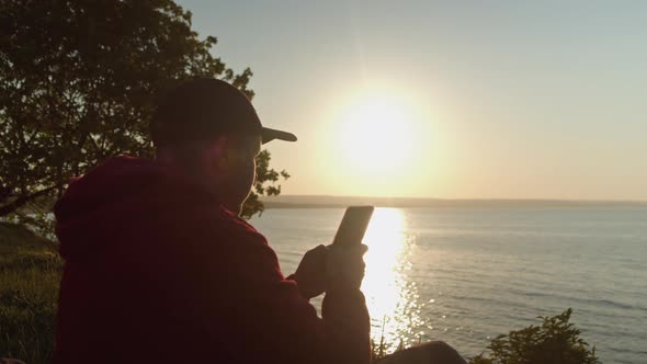 Man Traveler is Sitting on the Ocean or Sea Shore Using a Smartphone