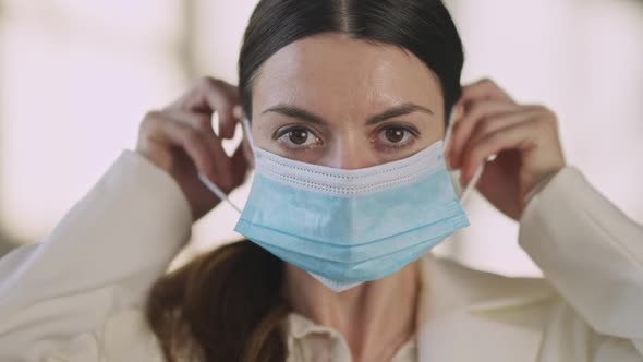 Headshot of Young Caucasian Woman with Big Brown Eyes Putting on Covid19 Face Mask Looking at Camera