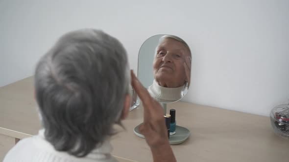 Adult gray-haired woman looks in the mirror. Stately mature woman with makeup straightens hair.