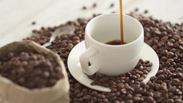 Drops Of Black Coffee Water Fall Into White Cup. White Porcelain Cup And Mug. Coffee Beans On Table