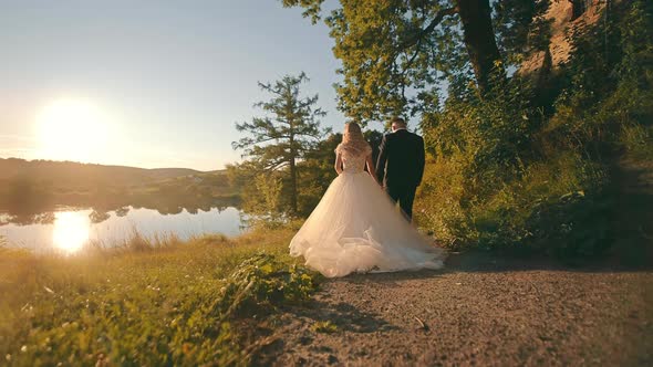 Happy Bride in White Dress and Groom Walking Down Country Road Holding Hands in Setting Sun