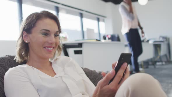 Caucasian businesswoman sitting in armchair, smiling and using smartphone