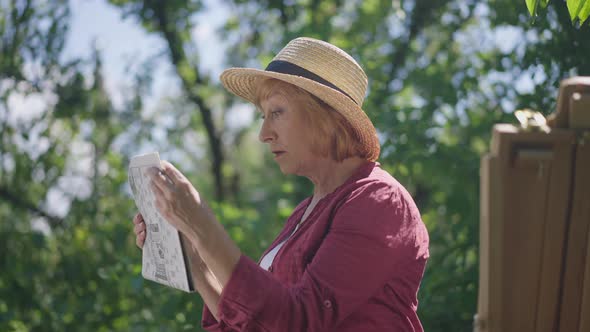 Side View Curios Senior Caucasian Woman Reading Newspaper Outdoors Standing in Garden with Easel