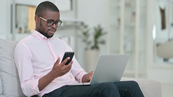 African Man Using Smartphone and Laptop at Home 