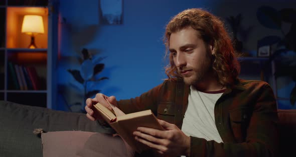 Smart Young Man Sitting on Sofa with Book and Looking Sleepy Late at Night, Handsome Hipster Guy