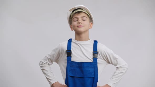 Caucasian Boy in Hard Hat and Overalls Putting Hands on Hips Posing at White Background