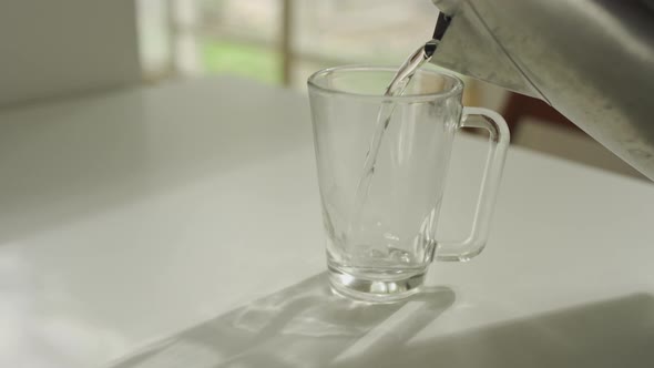 Hot water being poured into a glass mug from metal kettle in apartment