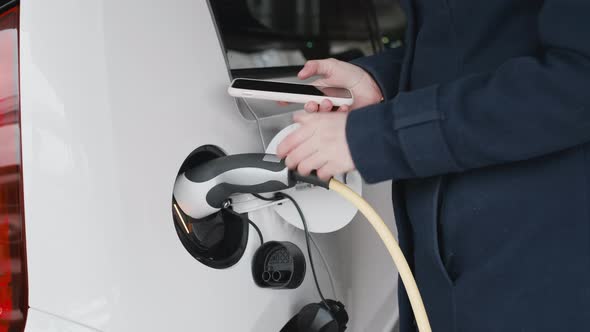 Woman Inserts the Power Supply Cable Into an Electric Car for Charging and Using Her Smartphone for