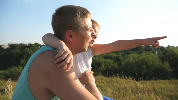 Redhaired Boy and His Father Sitting at Green Grass on the Lawn and Pointing at Something Outdoor