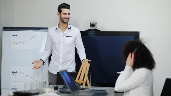 Portrait of Joyful Middle Eastern Man Dancing in Office with Caucasian Woman in Headphones Sitting