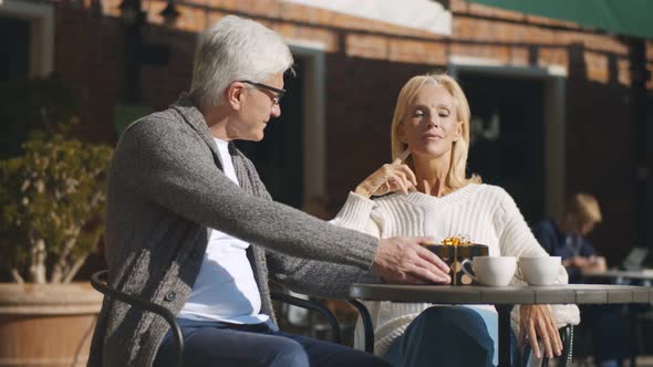 Senior Man Giving Nice Present To Wife Sitting Together on Restaurant Terrace