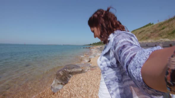 POV Shot Of Young Woman Reaching Down To Pick Up Plastic Bottle On Beach