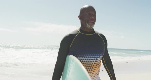 Senior african american man walking with surfboard on sunny beach