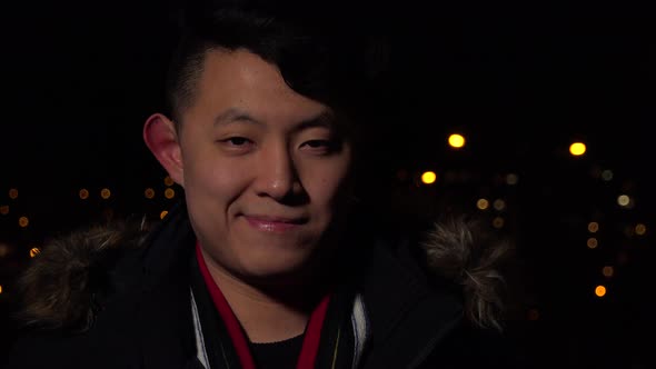 A Young Asian Man Smiles at the Camera in an Urban Area at Night - Face Closeup