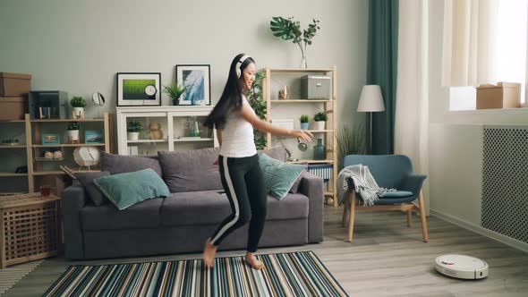 Cute Asian Female Student Is Listening To Music in Headphones and Dancing While Robotic Vacuum
