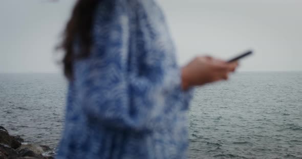 A Woman is Typing on a Mobile Phone While Standing on the Rocks on the Seashore