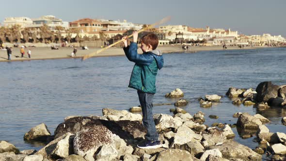 the Boy Spins a Stick with His Hands on the Rocks By the Sea