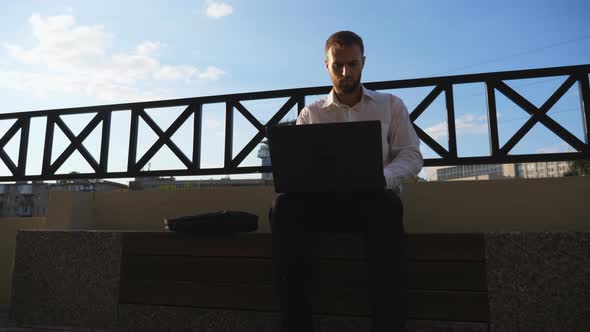 Young Employee Works on Notebook Sitting on Bench at City