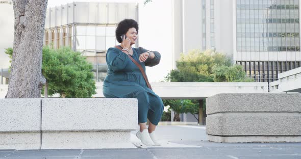 Happy plus size biracial woman talking on smartphone and checking smartwatch