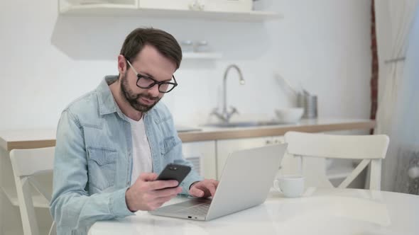 Beard Young Man Using Smartphone and Working on Laptop in Office 