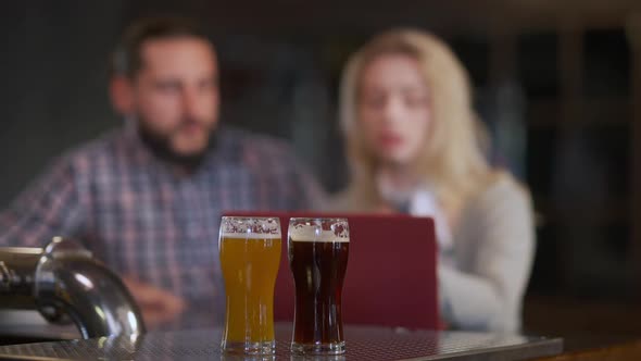 Glasses of Pale and Dark Lager Beer on Bar Counter with Blurred Couple of Freelancer Discussing