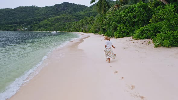 Camera Follows Young Woman As She Run on the Paradise Sandy Beach with Green Foliage and Shallow