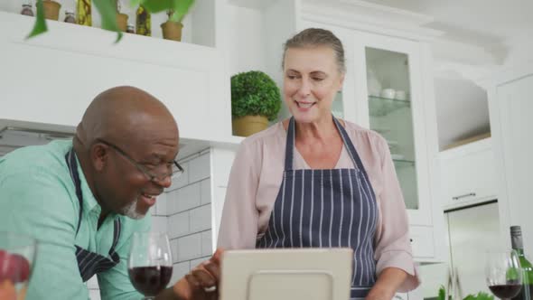 Smiling senior diverse couple wearing blue aprons and using tablet in kitchen