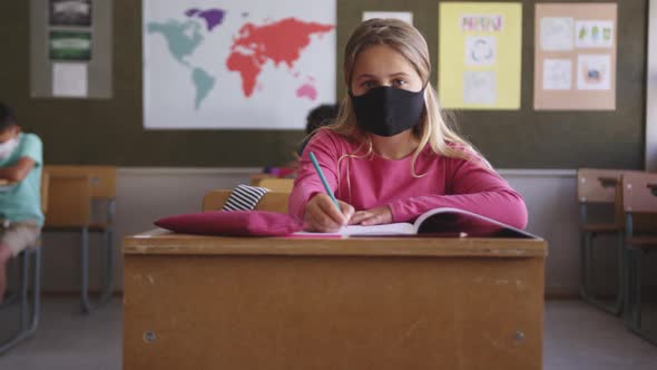 Girl wearing face mask writing while sitting on her desk at school 