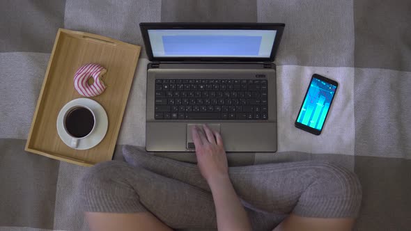 Overhead Shot, Girl Works Using a Laptop Sitting on a Bed, Eats a Donut and Drinks Coffee