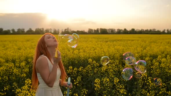 Young Redhair Woman Blowing Bubbles at the Camera Outdoors in Summer Meadow