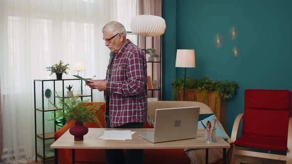Senior Grandfather Man Working on Laptop Computer Making Notes on Sheet of Paper Calculating Bills