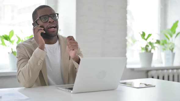 African Man Talking on Smartphone While Using Laptop in Office