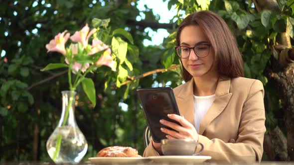 Young Business Woman Sits in a Cafe and Writes Text Messages on a Tablet. Girl with Coffee and a Bun