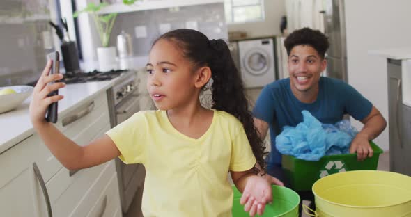 Happy biracial father and daughter making video call using smartphone, sorting waste together