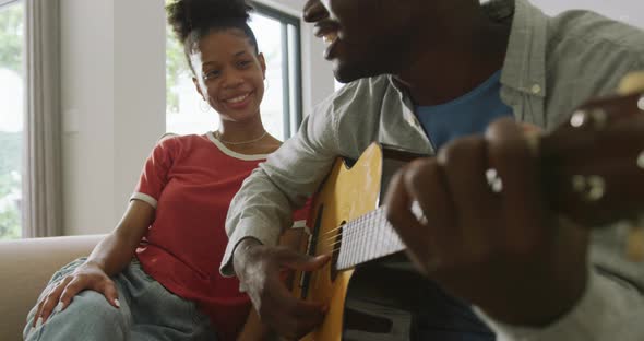 Video of happy african american couple sitting on sofa, singing and playing guitar