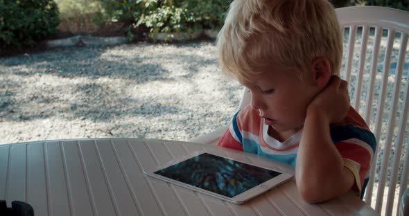 Little Tired Boy Learns Remotely From Home on a Tablet Sitting on the Terrace