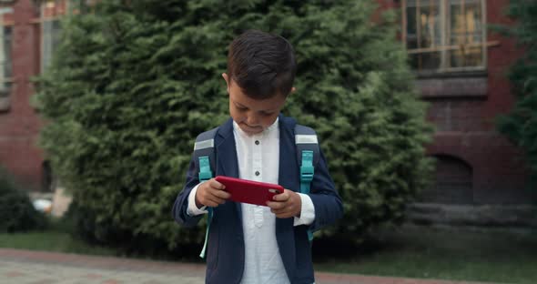 Boy Playing Online Game on Mobilephone While Walking at Street. Kid in School Uniform with Bag