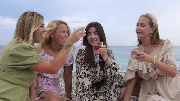 Four Women Clink Glasses of Wine at a Picnic By the Sea and Laughing