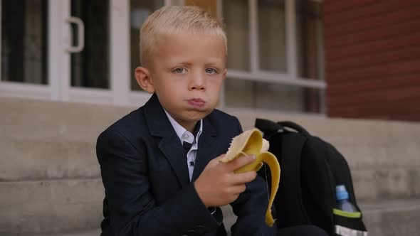 A Small Schoolboy Eats a Banana on the Steps of the School During Lunch