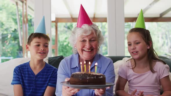 Caucasian senior woman in party hat blowing candles on birthday cake while her grandchildren sitting