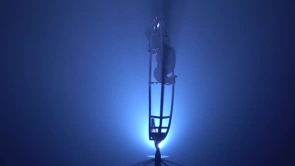 Gymnast Hangs Upside Down in a Twine on a Rotating Metal Construction . Blue Smoke Background