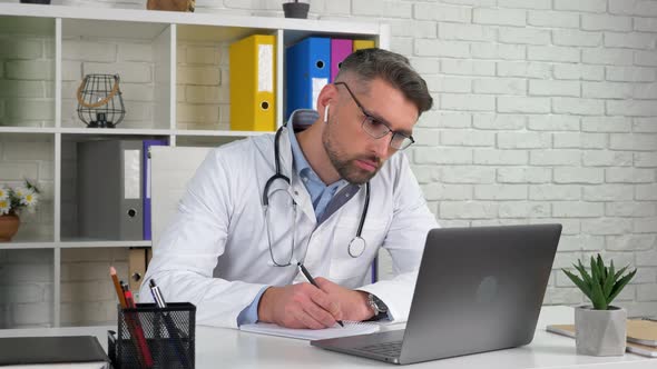 Doctor wears white medical coat in hospital office using laptop writes notebook