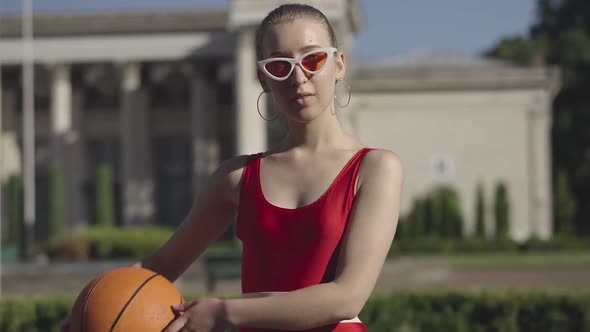 Portrait of Slim Confident Young Woman in Red Sportswear Posing with Basketball Ball Outdoors and