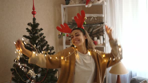 Young Cheerful Pretty Woman Wearing Reindeer Antlers Headband Dancing with Christmas Lights at Cozy