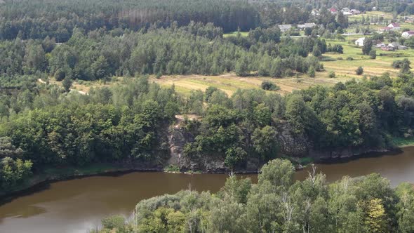 Aerial View of River Bordered by Forest Approaching Cliff With Field in Background