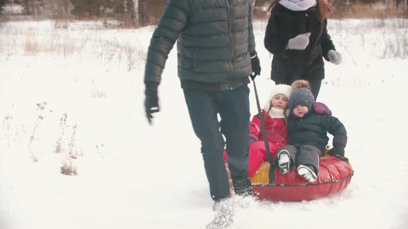 Family Running Outdoors Near the Forest - Man Rolls His Kids on the Inflatable Sled