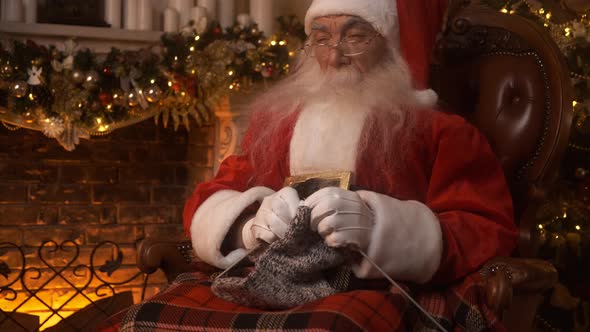 Santa Claus Holds a Knitted Sock in His Hands Near Fireplace